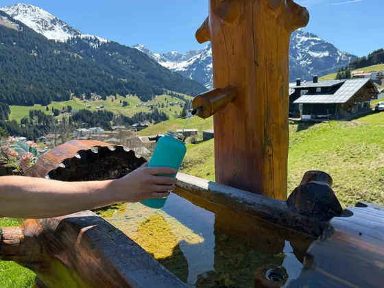 Hand filling a water bottle at the wooden fountain with fresh spring water against a mountain panorama in Hirschegg